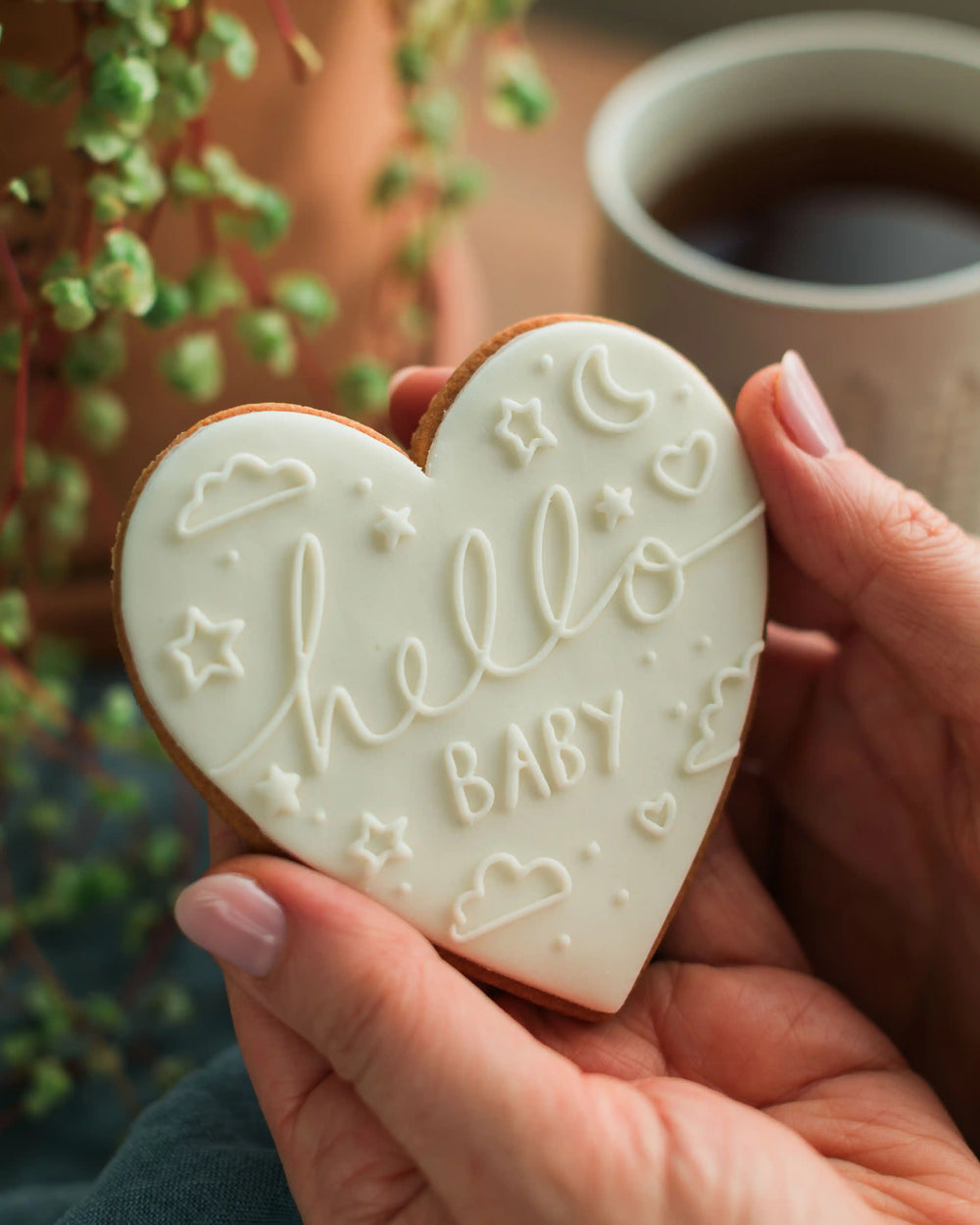 Honeywell Bakes Hello Baby Biscuit White - Heart-shaped cookie with 'hello baby' text held by a hand, with a cup of coffee in the background.