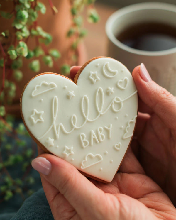 Honeywell Bakes Hello Baby Biscuit White - Heart-shaped cookie with 'hello baby' text held by a hand, with a cup of coffee in the background.
