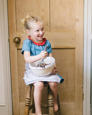 Honeywell Bakes Young Bakers Subscription, Young girl sitting on a stool in a kitchen, holding a bowl and whisk.