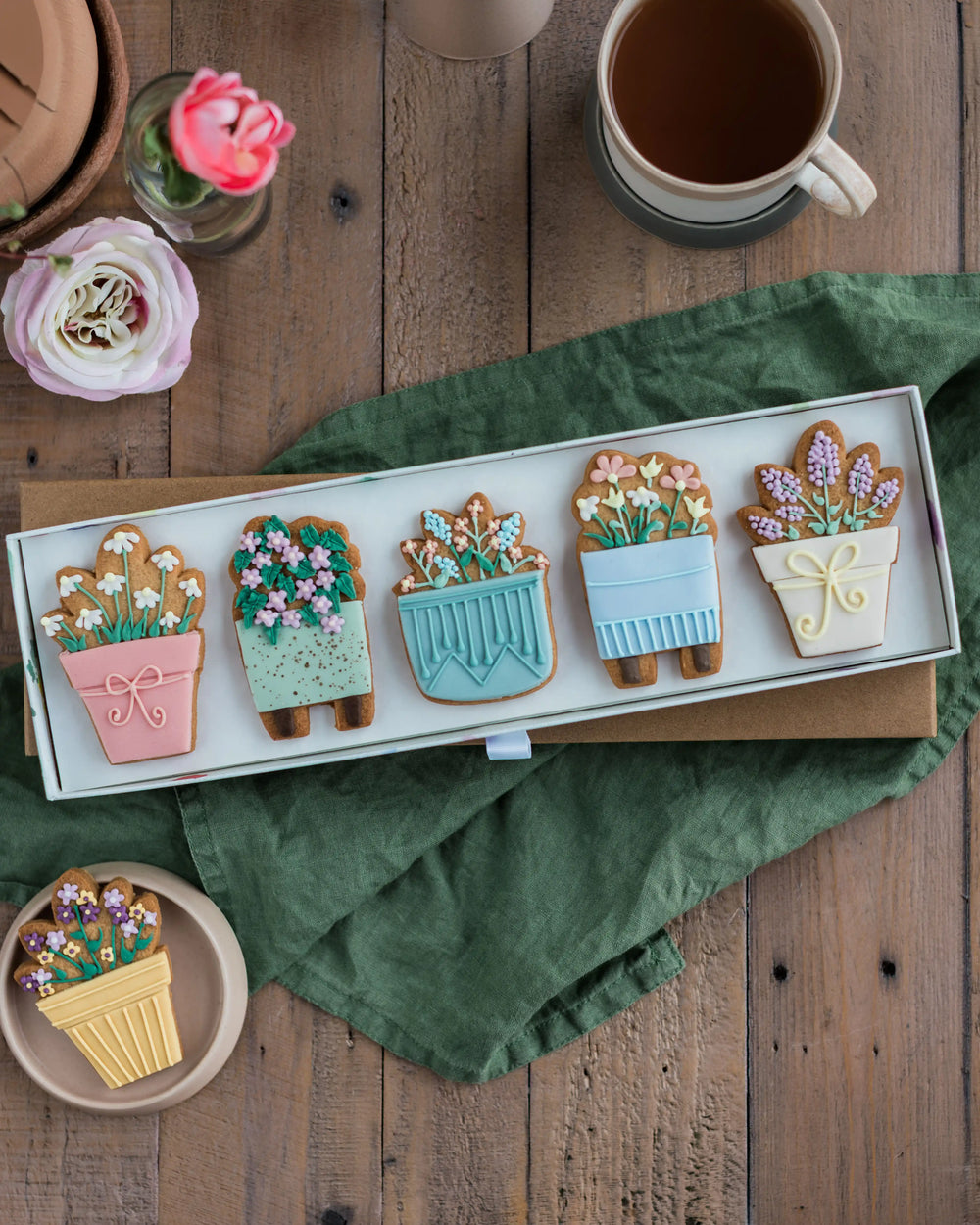 Honeywell Bakes House Plant Biscuit Gift Set - Decorative cookies shaped like potted plants on a wooden table with flowers and a cup of coffee.