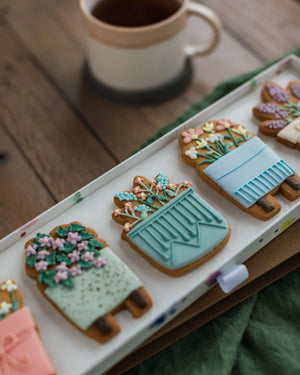 Honeywell Bakes House Plant Biscuit Gift Set - Decorative cookies shaped like potted plants on a tray with a cup of coffee in the background.