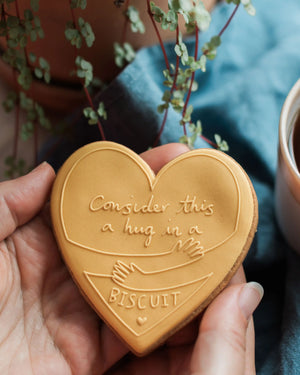 Honeywell Bakes Hug in a Biscuit - Heart-shaped biscuit with a message held by a hand, with a plant and cup in the background.