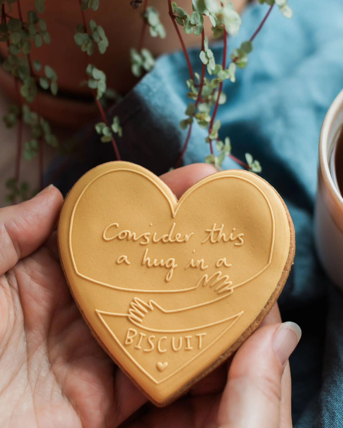 Honeywell Bakes Hug in a Biscuit - Heart-shaped biscuit with a message held by a hand, with a plant and cup in the background.