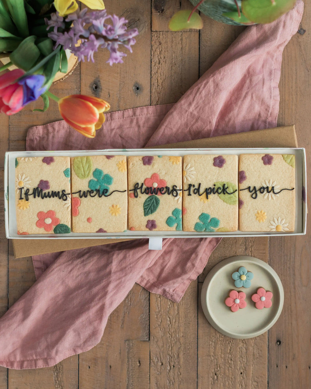 Honeywell Bakes If Mums Were Flowers Biscuit Gift Set - Box of cookies with floral designs and text on a wooden surface with flowers and a pink cloth.