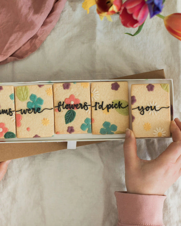 Honeywell Bakes If Mums Were Flowers Biscuit Gift Set - Hand holding a set of biscuits with floral designs and text on a textured surface with flowers in the background.