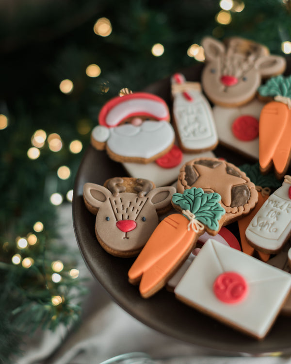 Honeywell Bakes Christmas Biscuits in a Jar - Decorated Christmas cookies shaped like Santa Claus, reindeer, carrots, and an envelope, arranged on a dark plate on Christmas Eve with festive blurred lights and greenery in the background.
