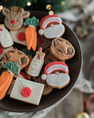 Honeywell Bakes Christmas Biscuits in a Jar - Decorated Christmas cookies shaped like Santa Claus, reindeer, carrots, and an envelope, arranged on a dark plate on Christmas Eve with festive blurred lights and greenery in the background.