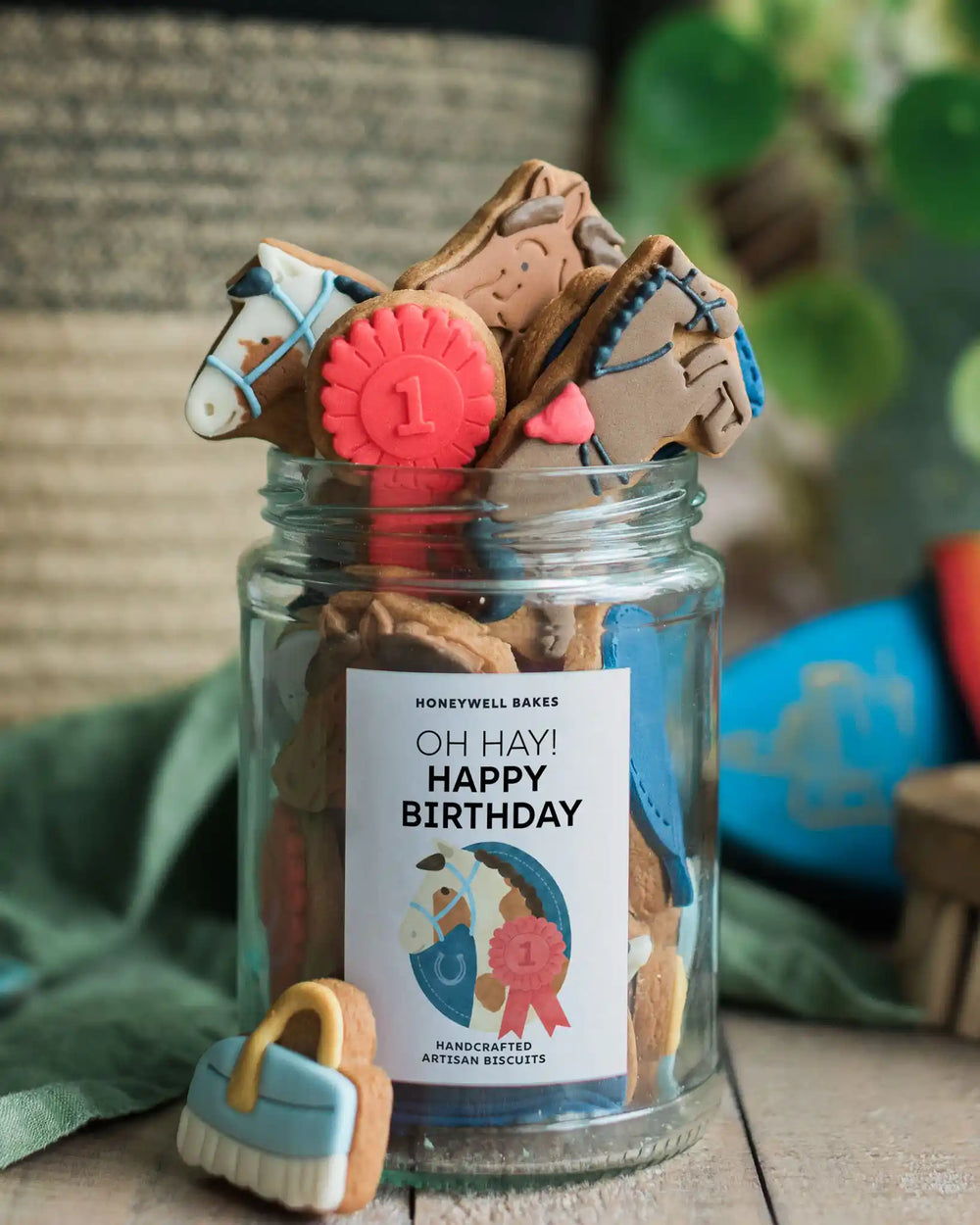 Honeywell Bakes - Jar of Horse Biscuits - A glass jar labeled 'Honeywell Bakes Jar of Horse Biscuits' filled with horse-shaped and horse-themed handcrafted biscuits, with a small blue-and-white brush in front on a rustic tabletop.