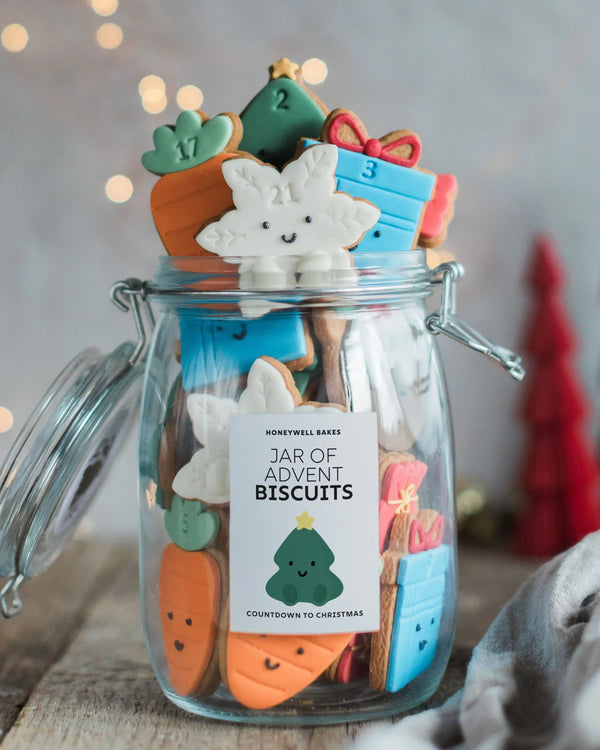 Honeywell Bakes Jar of Advent Biscuits - A clear glass jar filled with assorted Christmas-themed biscuits shaped like a carrot, tree, and gift boxes, with a white label reading 'Honeywell Bakes Jar of Advent Biscuits.' The jar is photographed on a rustic surface with a softly blurred holiday background and warm lights.