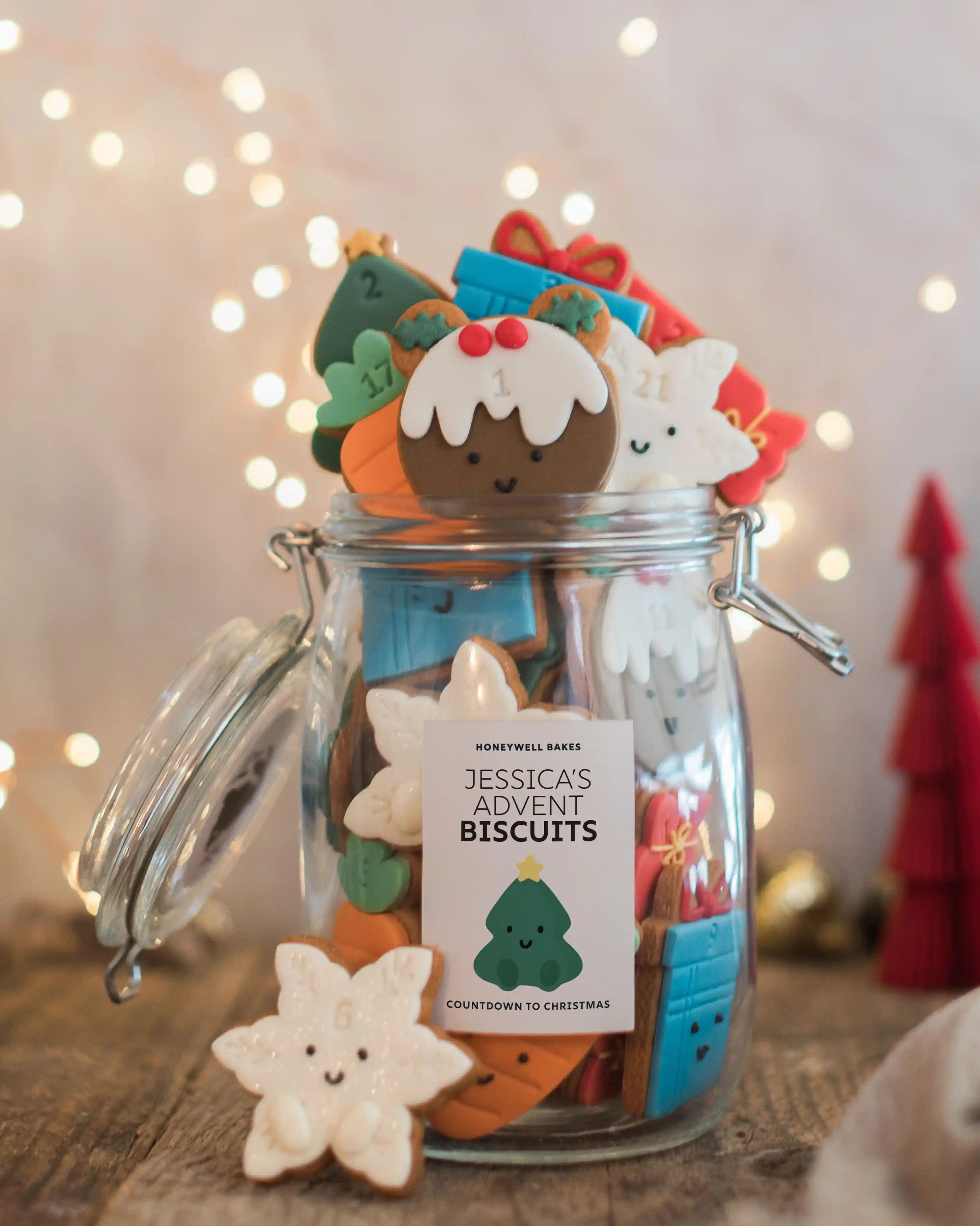 Honeywell Bakes Jar of Advent Biscuits - A glass jar filled with colourful Christmas-themed biscuits, including a gingerbread man, carrot, reindeer, and star-shaped cookies, with a label reading 'Jessica's Advent Biscuits'. The jar is set on a wooden surface with warm bokeh string lights in the background.