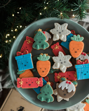 Honeywell Bakes Jar of Advent Biscuits - A round light blue plate filled with decorated Christmas sugar cookies shaped like carrots, snowflakes, gingerbread houses, gifts, and a tree, set against a festive background with a Christmas tree and warm lights.