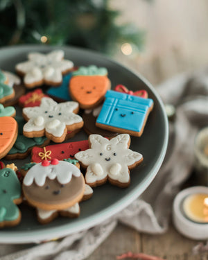 Honeywell Bakes Jar of Advent Biscuits - A plate of decorated Christmas cookies shaped like snowflakes, carrots, a gift box, and a gingerbread figure, styled on a rustic table with a candle and soft holiday background.