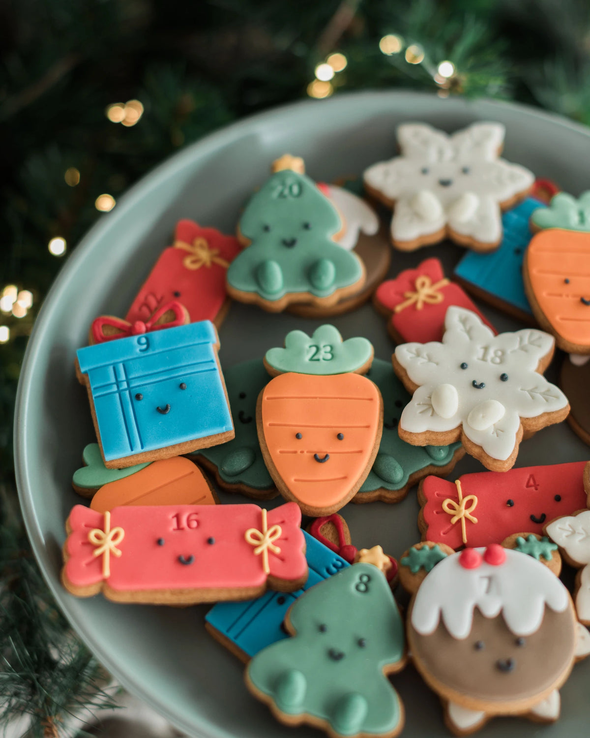 Honeywell Bakes Jar of Advent Biscuits - A plate of decorated Christmas sugar cookies shaped like a carrot, snowflake, gift box, and Christmas tree, arranged on a gray plate with a blurred festive background.
