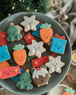 Honeywell Bakes Jar of Advent Biscuits - A plate of decorated Christmas advent cookies shaped like carrots, snowflakes, gifts, and a tree, arranged on a gray plate with festive holiday decor in the background.