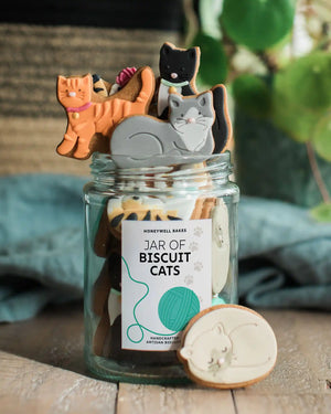 A glass jar filled with cat-shaped artisan biscuits, with several cookies sticking out of the top and one biscuit placed in front. The jar label reads 'HONEYWELL BAKES JAR OF BISCUIT CATS.