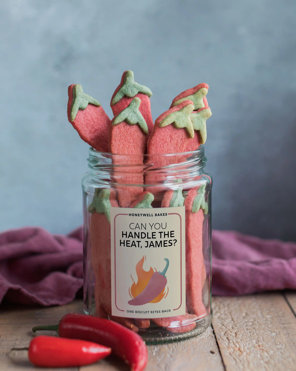 Honeywell Bakes Jar of Chilli Roulette Biscuits filled with red chilli-shaped cookies topped with green leafy stems, with two whole red chilli peppers in front. The jar has a label that reads ‘CHILLI BISCUIT ROULETTE'.