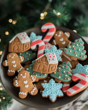 Honeywell Bakes Jar of Gingerbread Men Biscuits - A dark plate filled with decorated Christmas gingerbread cookies, including gingerbread men, houses, candy canes, snowflakes, and trees, set against a blurred festive background with warm lights and greenery.