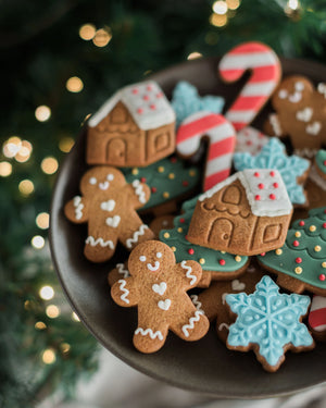 Honeywell Bakes Jar of Gingerbread Men Biscuits - A dark plate filled with decorated Christmas gingerbread cookies, including gingerbread men, houses, candy canes, snowflakes, and trees, set against a blurred festive background with warm lights and greenery.