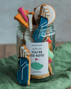 Honeywell Bakes Jar of Golf Biscuit Gift - Jar of cookies shaped like golf clubs and balls with a decorative label on a green surface.