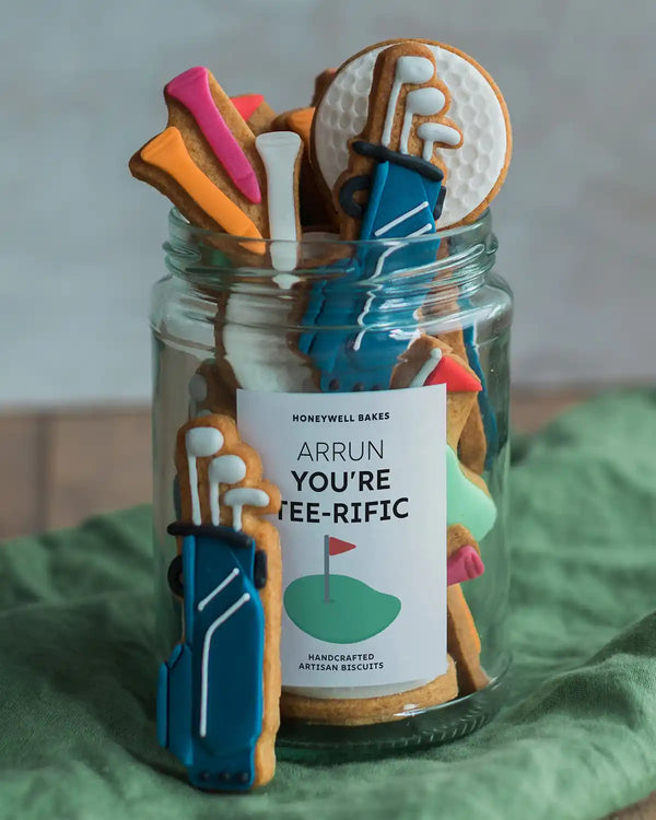Honeywell Bakes Jar of Golf Biscuit Gift - Jar of cookies shaped like golf clubs and balls with a decorative label on a green surface.