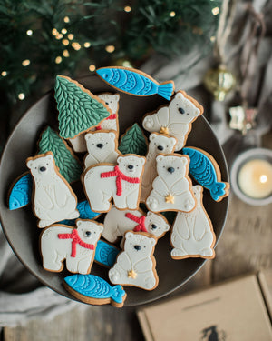 Honeywell Bakes Jar of Polar Bear Biscuits - A top-down view of decorated Christmas sugar cookies shaped like polar bears, fish, and pine trees, arranged on a dark plate with a festive holiday background.