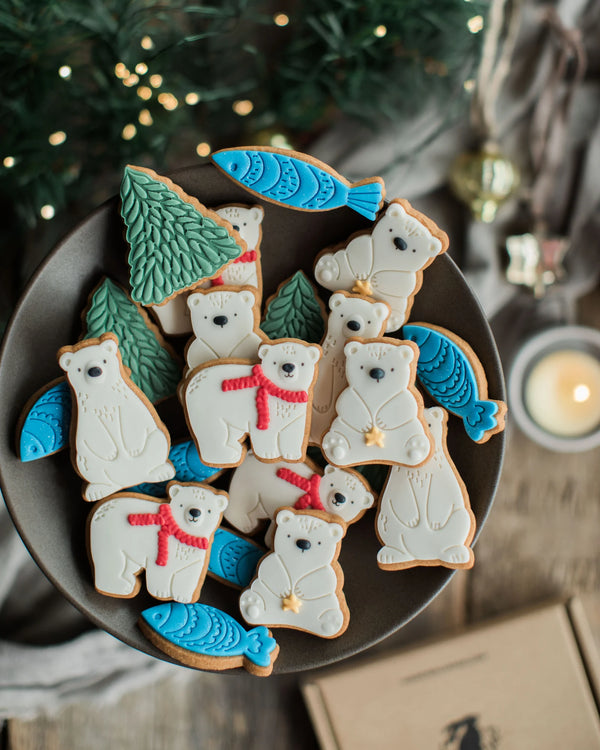 Honeywell Bakes Jar of Polar Bear Biscuits - A top-down view of decorated Christmas sugar cookies shaped like polar bears, fish, and pine trees, arranged on a dark plate with a festive holiday background.