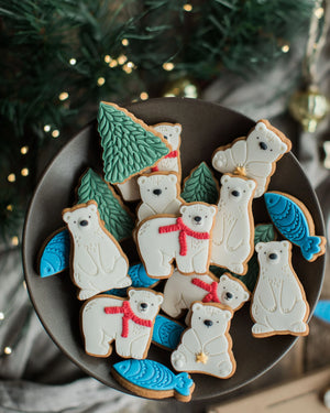 Honeywell Bakes Jar of Polar Bear Biscuits - A top-down view of decorated polar bear and fish shaped sugar cookies arranged on a dark round plate, with green pine trees and warm blurred holiday lights in the background.