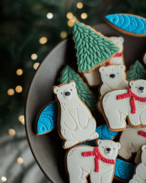 Honeywell Bakes Jar of Polar Bear Biscuits - Decorated polar bear and tree-shaped cookies arranged on a dark plate, with blurred holiday lights and greenery in the background.