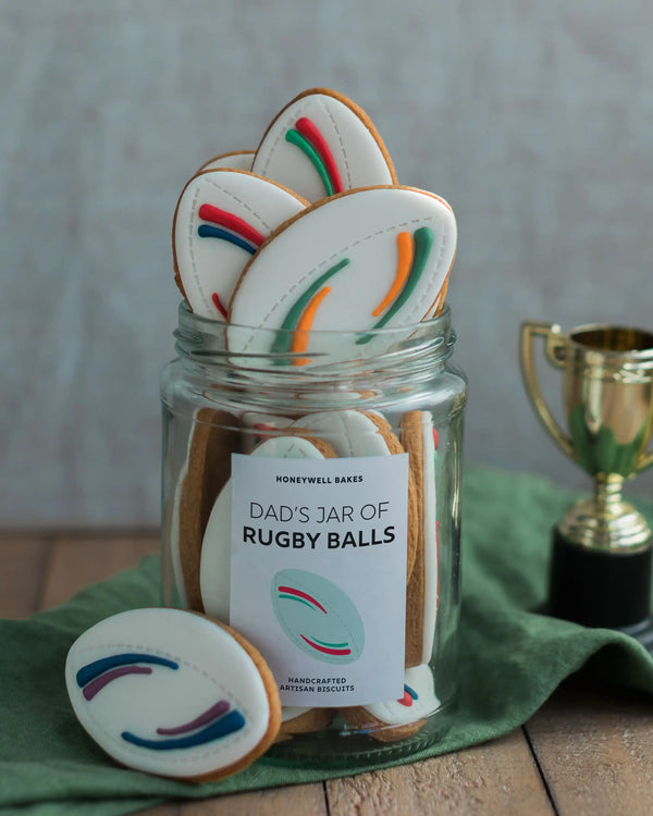 Honeywell Bakes Jar of Rugby Balls Biscuit Gift - Jar labeled 'Dad's Jar of Rugby Balls' filled with cookies shaped like rugby balls, on a wooden surface with a green cloth and trophy.