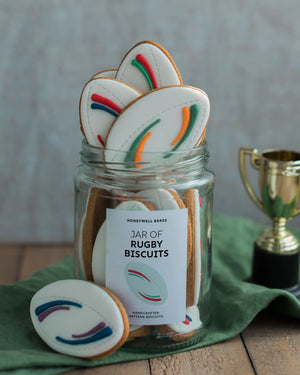 Honeywell Bakes Jar of Rugby Balls Biscuit Gift - Jar of rugby-themed cookies with a decorative label, surrounded by cookies on a wooden surface.