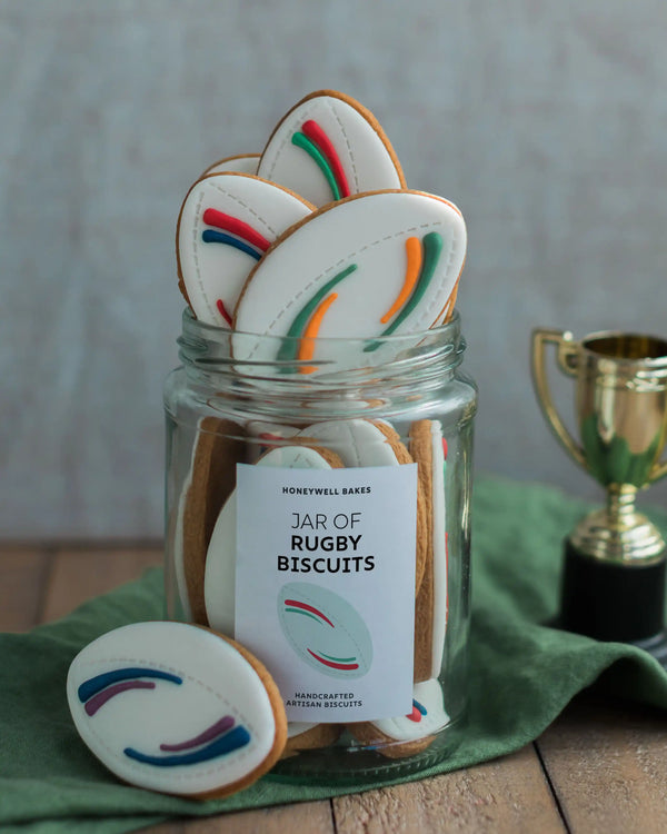 Honeywell Bakes Jar of Rugby Balls Biscuit Gift - Jar of rugby-themed cookies with a decorative label, surrounded by cookies on a wooden surface.