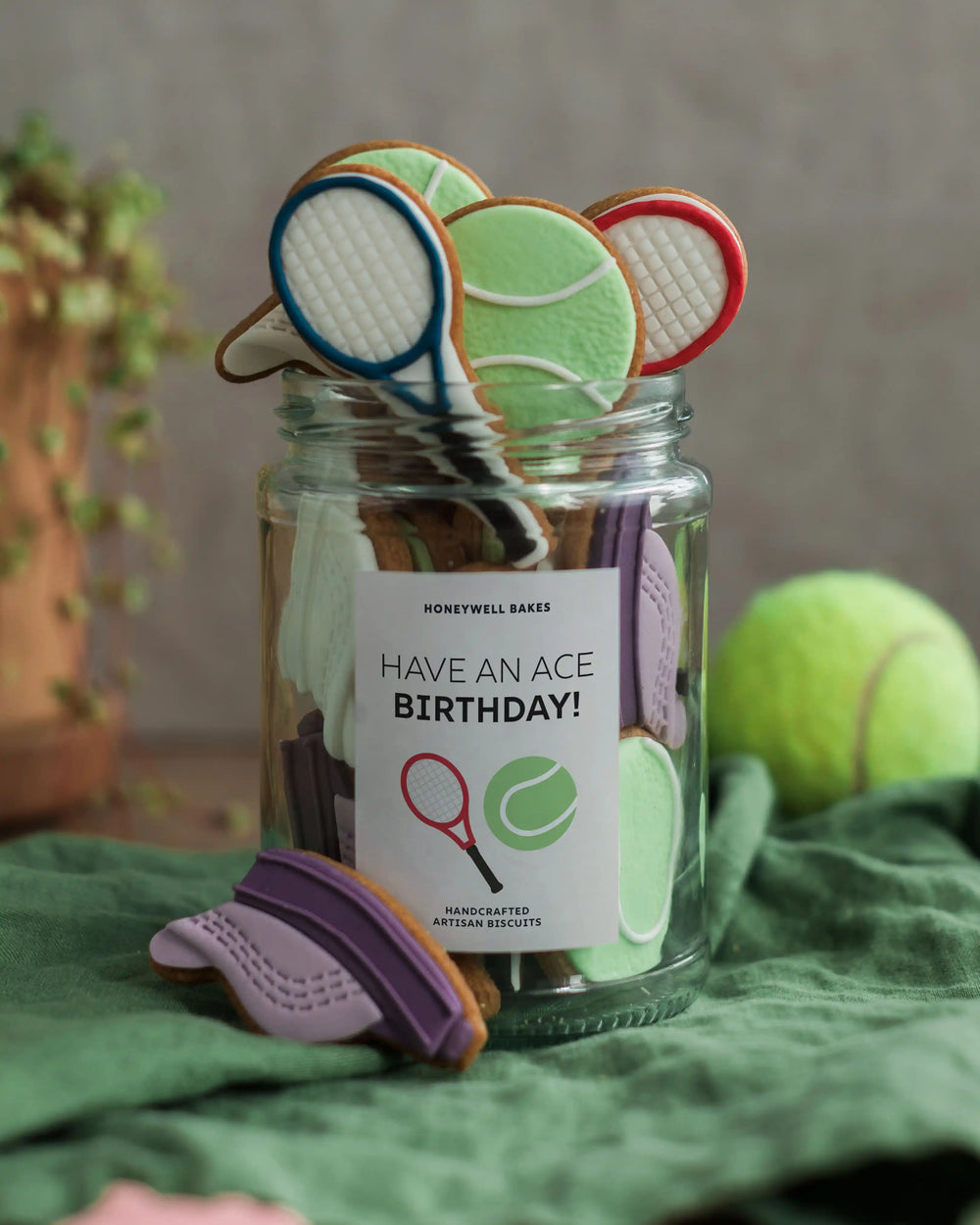 Honeywell Bakes Jar of Tennis Biscuit Gift - Jar of cookies shaped like tennis rackets and balls with a 'Have an Ace Birthday!' label on a green surface.
