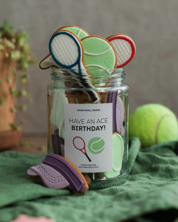 Honeywell Bakes Jar of Tennis Biscuit Gift - Jar of cookies shaped like tennis rackets and balls with a 'Have an Ace Birthday!' label on a green surface.