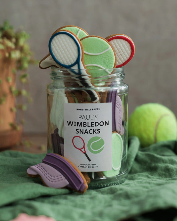 Honeywell Bakes Jar of Tennis Biscuit Gift - Jar of cookies shaped like tennis rackets and balls with a 'Paul's Wimbledon Snacks' label, on a green surface.