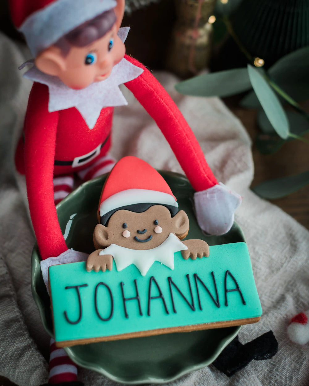 A close-up of a Jolly The Elf - Personalised Biscuit with the name 'JOHANNA' in black icing on a turquoise base, held by a red elf doll in a green bowl on a neutral fabric surface.