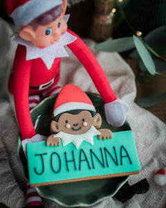 A close-up of a Jolly The Elf - Personalised Biscuit with the name 'JOHANNA' in black icing on a turquoise base, held by a red elf doll in a green bowl on a neutral fabric surface.