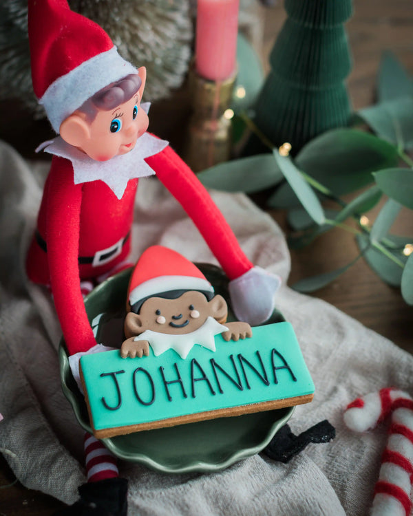 A close-up of a Jolly The Elf - Personalised Biscuit with the name 'JOHANNA' in black icing on a turquoise base, held by a red elf doll in a green bowl on a neutral fabric surface.