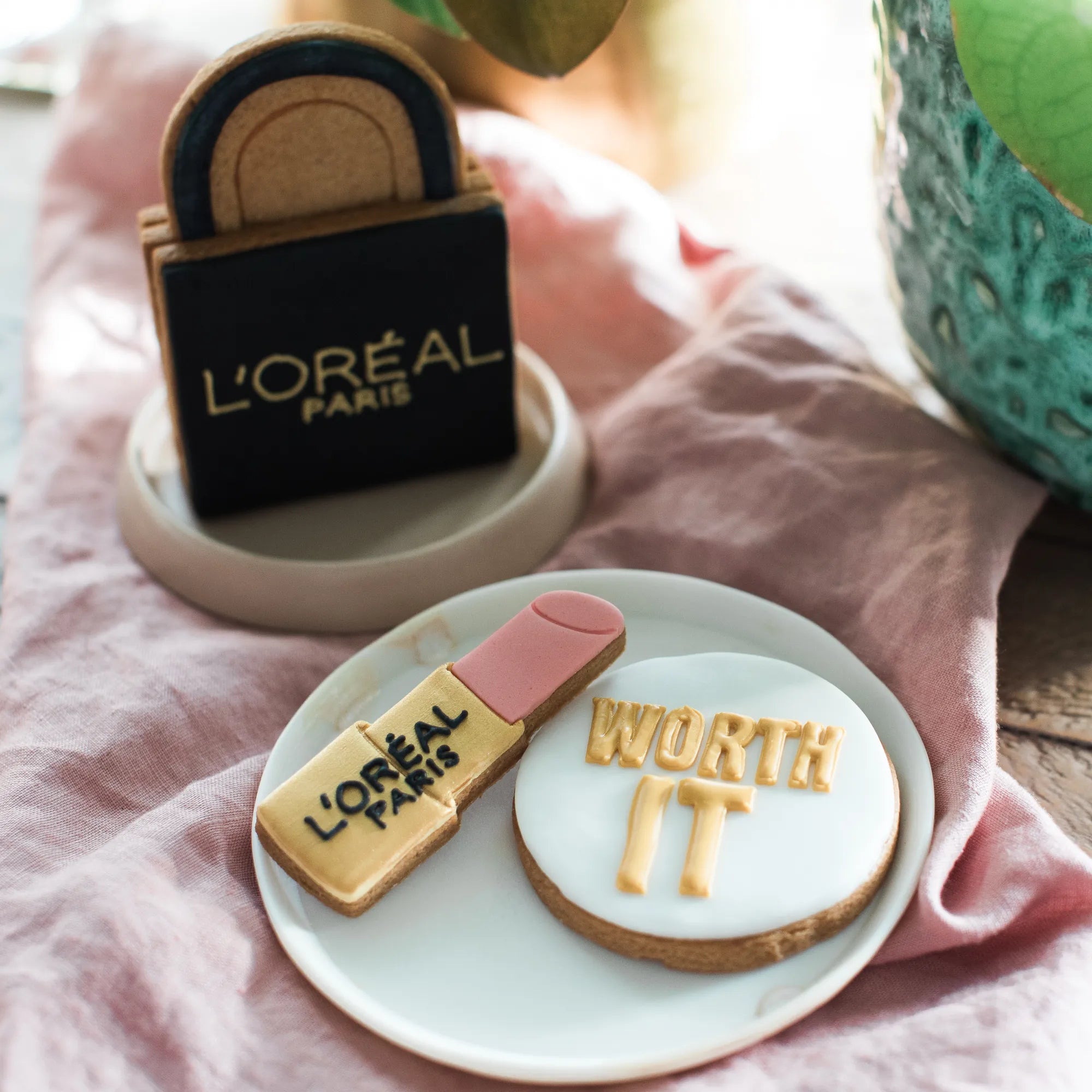 Honeywell Bakes  L'Oréal Paris themed cookies; one cookie is shaped like a lipstick tube and the other is round with white icing and gold lettering reading 'WORTH IT'. There is a 3D L'oreal biscuit retail bag.