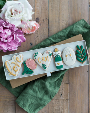 Honeywell Bakes Large Lovely Couple Biscuit Gift Set - Decorative cookies with floral and bottle designs on a wooden surface with flowers.