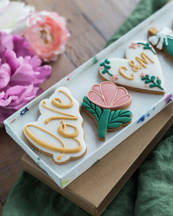 Honeywell Bakes Large Lovely Couple Biscuit Gift Set - Decorative cookies with floral designs and 'LOVE' text on a white tray with flowers in the background.