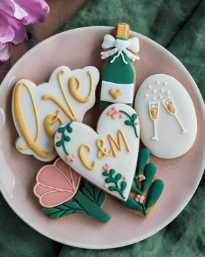 Honeywell Bakes Large Lovely Couple Biscuit Gift Set - Decorative cookies shaped like a bottle, heart, and flowers on a pink plate with a green background.