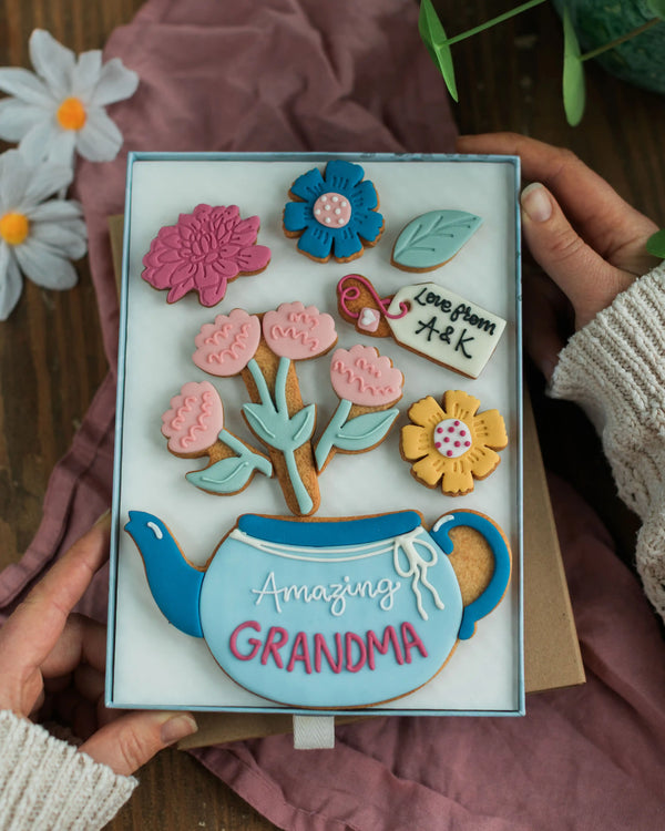 Honeywell Bakes Large Teapot Blooms Biscuit Gift - Decorative cookie box with floral designs and 'Amazing Grandma' text, held by hands on a wooden surface.