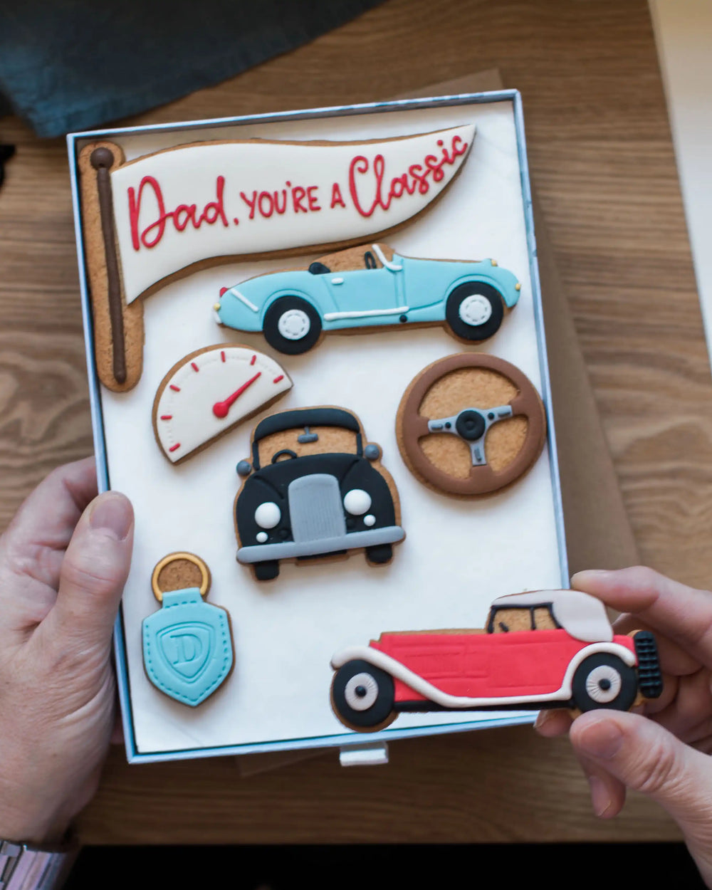 Honeywell Bakes Large You’re A Classic Biscuit Gift - Cookie set with car-themed decorations in a box, held by two hands on a wooden surface.