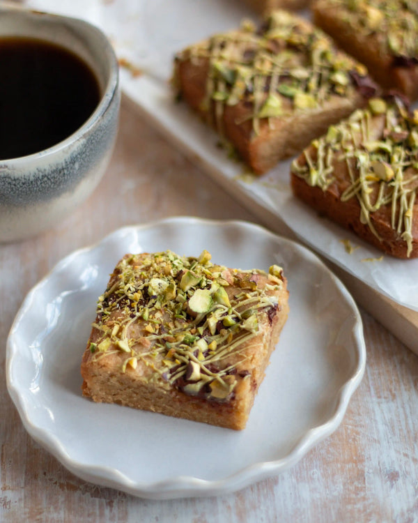 Honeywell Bakes Limited Edition Brown Butter Pistachio Blondies - squares of blondies with a pistachio and chocolate topping, on a wooden board with a cup of coffee in the background