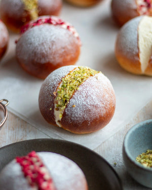 Honeywell Bakes Limited Edition Lemon & Raspberry Maritozzi Buns - large bread bun filled with cream and topped with pistachios, on a wooden table with more buns in the background
