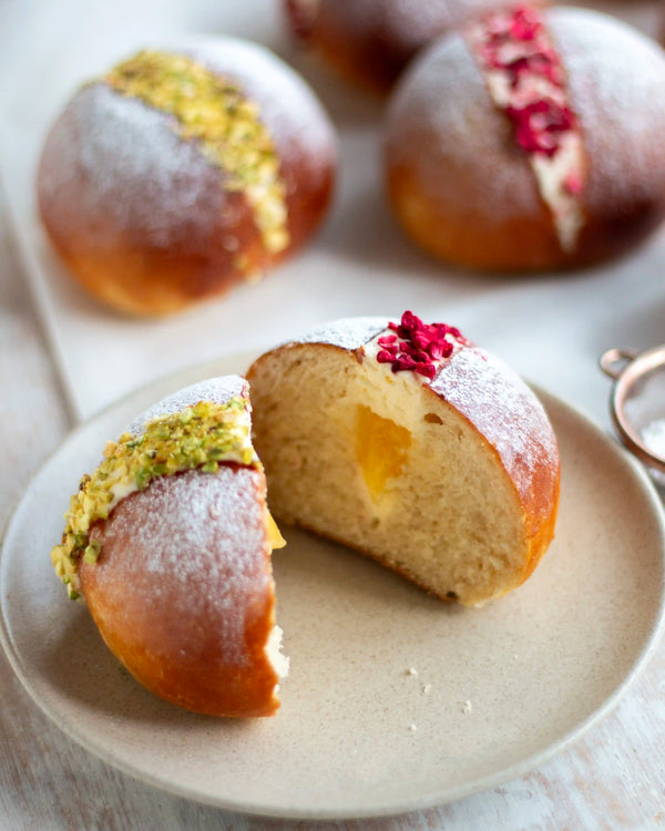 Honeywell Bakes Limited Edition Lemon & Raspberry Maritozzi Buns - large bread bun cut open and filled with cream and  lemon curd topped with pistachios and raspberries, on a wooden table with more buns in the background