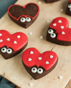 Honeywell Bakes Love Bug Biscuit Baking Kit - Heart-shaped cookies with red icing, chocolate bodies, and white candy eyes on a wooden surface.