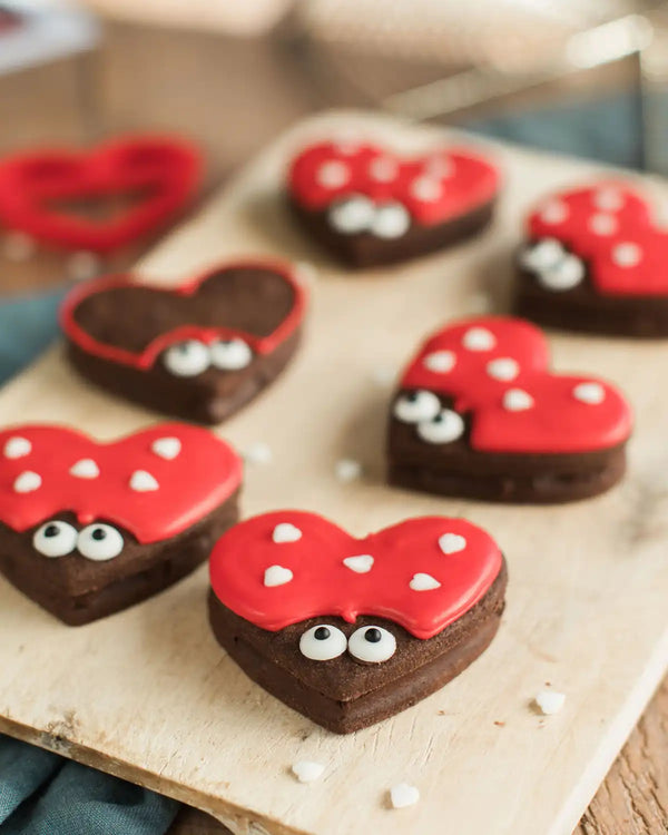 Honeywell Bakes Love Bug Biscuit Baking Kit - Heart-shaped cookies with red icing and white sprinkles on a wooden board.