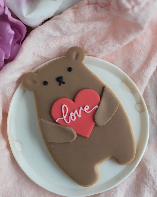 Honeywell Bakes Love Heart Biscuit Bear - Cookie shaped like a bear holding a heart with 'love' written on it, placed on a white plate with a pink floral background.