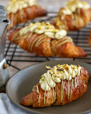 Honeywell Bakes Make Your Own Pistachio Croissant Baking Kit - Croissants with pistachio buttercream and pistachio drizzle on a plate, with more croissants on a cooling rack in the background.
